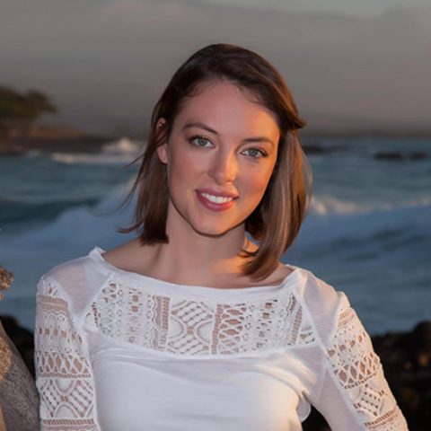 A smiling woman with shoulder-length brown hair at the beach during sunset, ocean waves in backgroun.